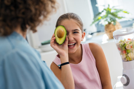 Shot of happy little girl having fun with her mother holding avocado slice at her face while posing...