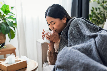 Shot of illness young woman drinking tea while lying with a blanket on the couch at homeの写真素材
