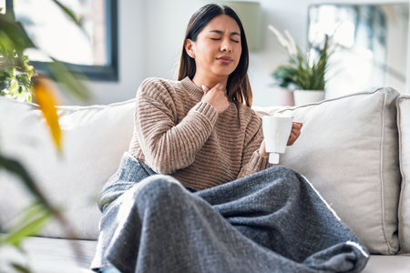 Shot of illness young woman with sore throat drinking tea while lying with a blanket on the couch at homeの写真素材