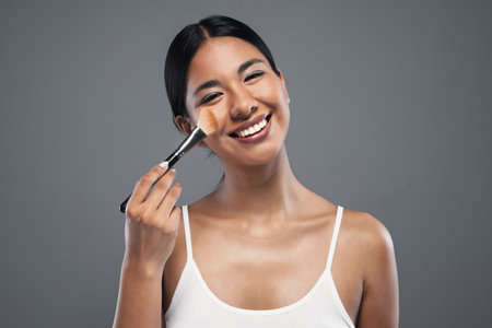 Shot of beautiful young woman doing make-up while looking at camera in isolated grayの写真素材