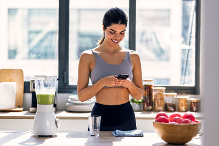 Shot of sporty young woman making a vegetable green smoothie while using smartphone in the kitchen at home.の写真素材
