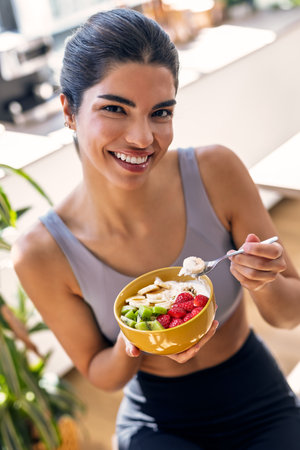 Portrait of smiling sporty woman eating a healthy fruit bowl while looking at camera sitting in the kitchen at homeの写真素材