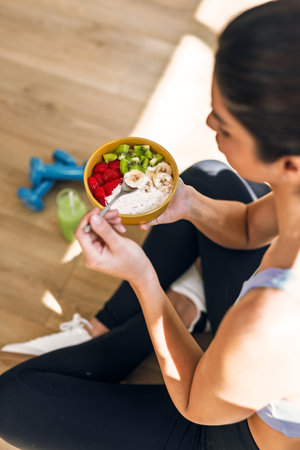 Close up of smiling sporty woman eating a healthy fruit bowl while sitting indoorsの写真素材
