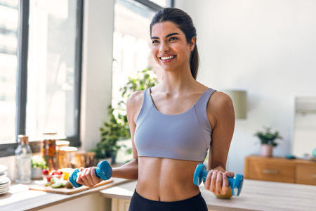 Shot of beautiful sporty woman working out with weights in the kitchen at homeの写真素材