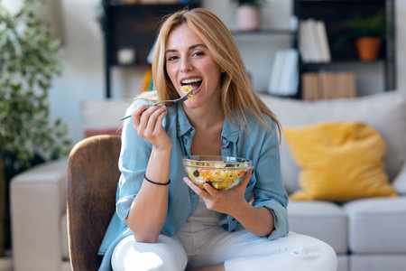 Portrait of smiling woman eating healthy pasta salad while sitting on chair looking at camera the living roomの写真素材