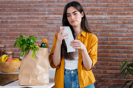 Shot of beautiful woman checking shopping ticket while leaving cardboard bag with healthy shopping...