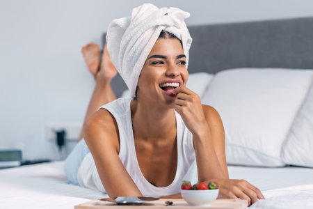Shot of smiling woman having strawberries for breakfast after taking a shower while lying on the bedの写真素材