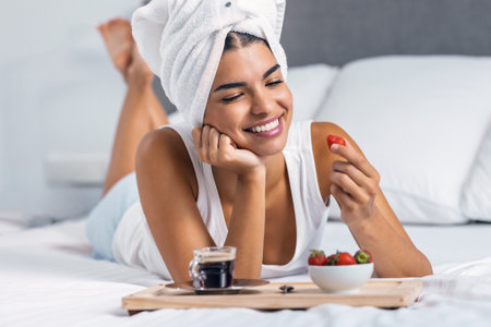 Shot of smiling woman having strawberries for breakfast after taking a shower while lying on the bedの写真素材