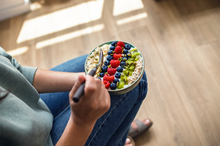 Close up of young woman eating a healthy fruit bowl in the kitchen at homeの写真素材