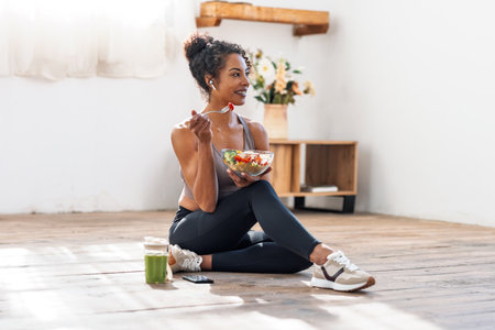 Shot of athletic woman eating a healthy fruit bowl while sitting on the floor at homeの写真素材
