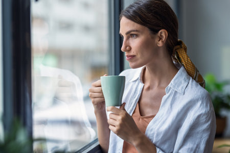 Shot of beautiful woman drinking a cup of coffee while looking forward standing next to the window at home.の写真素材