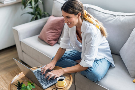 Shot of attractive woman working with laptop while drinking coffee sitting on the couch at homeの写真素材