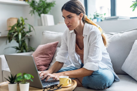 Shot of attractive woman working with laptop while drinking coffee sitting on the couch at homeの写真素材