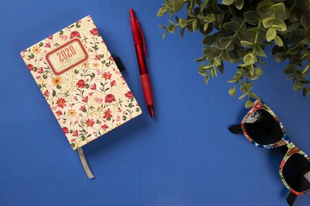 Blue office desk with a colorful diary, sunglasses and a green plant. Top view conceptの写真素材