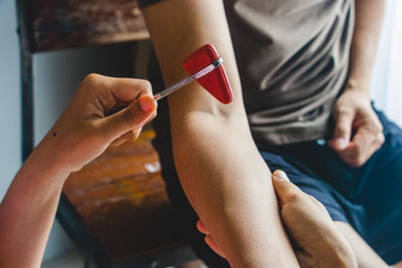 Doctor testing biceps tendon reflex with red hummer jerk on young man arm.の写真素材