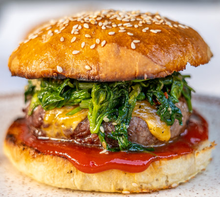 cheeseburger with beef patty, spinach, red sauce, cheddar cheese piled up in a self-made bun with sesameの写真素材