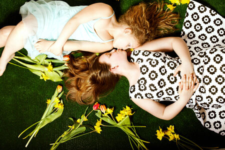 Two pretty young sisters lying over colorful flowers, kissing. Children's day. Happy family.の写真素材