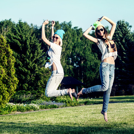 Two young beautiful girls jumping in park, wearing fashionable clothes. Summer photo. Best friends.の写真素材