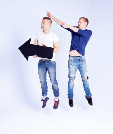 Two young fashionable man posing in studio with black empty arrow, jumping, smiling. Conceptual photo.の写真素材