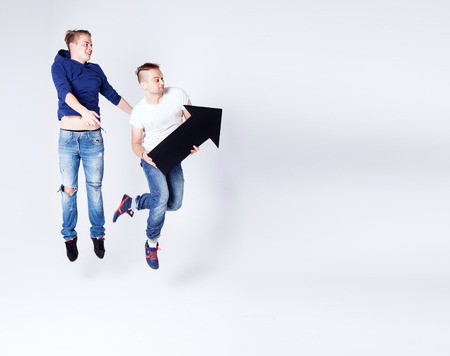 Two young fashionable man posing in studio with black empty arrow, jumping, smiling. Conceptual...