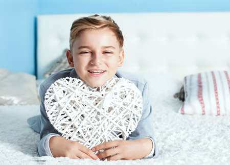 Handsome young teenage boy lying in bed, smiling, holding bog white heart and looking at camera.の写真素材