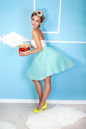 Beautiful blonde smiling woman holding box of strawberries . Indoor photo. Girl wearing fashionable dress, looking at cameraの写真素材