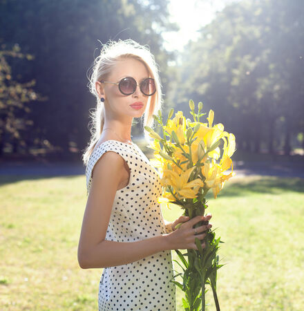 Beautiful young blonde woman posing with flowers on sunny day, wearing sunglassesの写真素材