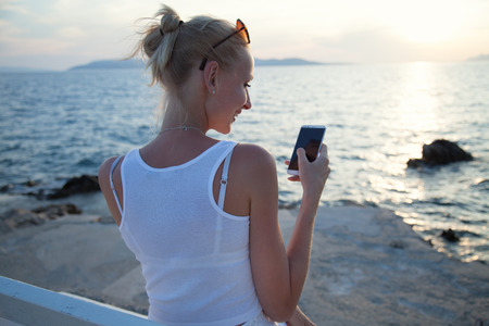 Beautiful young blonde woman using mobile phone, sitting on bench. Summer photo. Coastal view.