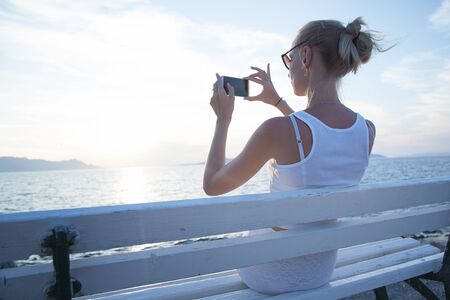 Beautiful young blonde woman using mobile phone, sitting on bench. Summer photo. Coastal view.の写真素材