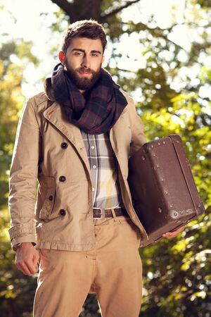 Fashion photo of young handsome man with suitcases in park. Autumn. Men wearing coat and scarf. Sunny day.の写真素材