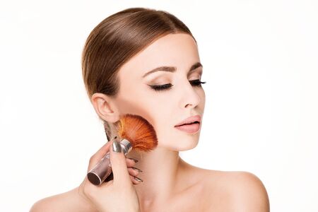 Beauty portrait of young natural woman holding brush in hand and applying cosmetic on face. Studio...