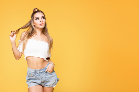 Young beautiful caucasian woman posing on yellow background. Girl in short jeans and white fashionable top. Studio shot.の写真素材