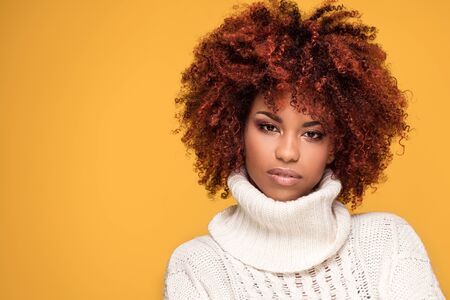 Young beautiful african american girl with an afro hairstyle. Attractive girl wearing cozy sweater. Portrait. Yellow background. Girl with glamour makeup.の写真素材