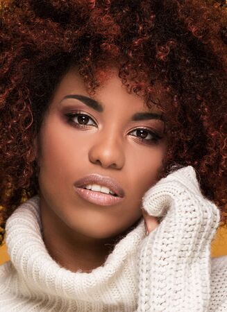 Young beautiful african american girl with an afro hairstyle. Attractive girl wearing cozy sweater. Portrait. Yellow background. Girl with glamour makeup.の写真素材