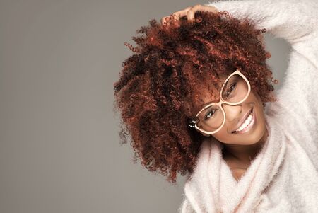 Happy beautiful african american girl with afro hairstyle posing in cozy sweater and eyeglasses, smiling.の写真素材