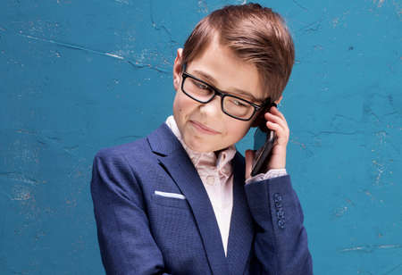 Kid in eyeglasses posing on blue background, talking by mobile phone. Child Boy smiling, elegant little businessman in fashionable clothes.の写真素材