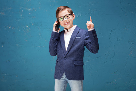 Kid in eyeglasses posing on blue background, talking by mobile phone. Child Boy smiling, elegant little businessman in fashionable clothes.の写真素材