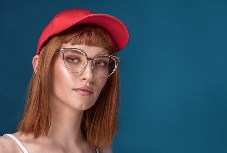 Portrait of attractive young redhead woman with fashionable eyeglasses and cap. Girl looking at camera. Blue background.の写真素材