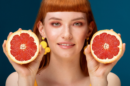 Portrait of beautiful redhead young girl holding two cutted grapefruit parts near her face before blue background. Natural woman with fashionable haircut and freckles on her face. Healthy freshness concept.の写真素材