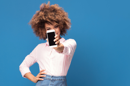 Beautiful afro woman showing mobile phone and smiling, standing on blue background. Girl wearing jeans short skirt and elegant shirt in white and pink stripes.の写真素材