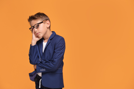 Little sad boy posing on orange studio backgroud, wearing fashionable eyeglasses and jacket.の写真素材