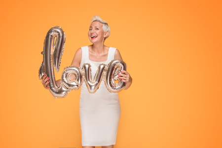 Attractive middle aged woman holding gold love balloon, smiling to the camera. Yellow studio background.の写真素材