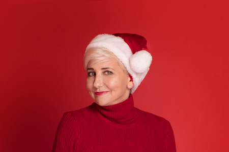 Happy smiling mature woman in red santa claus hat on red studio background.の写真素材
