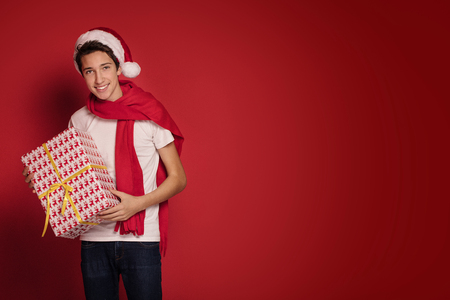 Happy teenager boy with Christmas gift posing in Santa Claus hat, smiling, red studio background.の写真素材