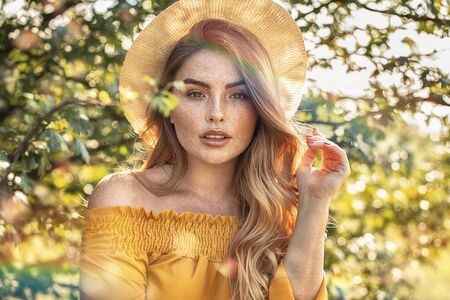 Happy beautiful woman with freckles looking to the camera, posing in hat in blooming garden at sunny day.の写真素材