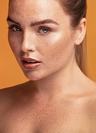 Closeup beauty portrait of girl with freckles on her face and body. Natural makeup. Ginger woman posing on orange studio background.の写真素材
