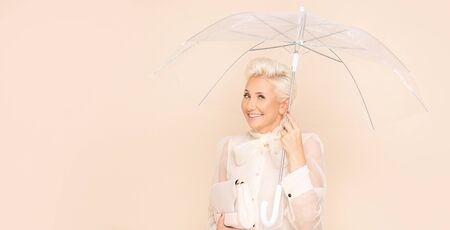 Fashion portrait of elegant adult woman with short blond hair posing in shirt with bow, holding handbag and umbrella, smiling to the camera. Middle age lady in glamour makeup.の写真素材