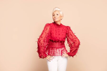 Fashion portrait of adult woman with short blond hair posing in red elegant shirt over beige background in studio. Middle age lady in glamour makeup.の写真素材
