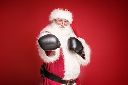 Real Santa Claus in boxing gloves is ready to fight , looking at camera, standing on red studio background. Merry Christmas and Happy New Year concept. の写真素材