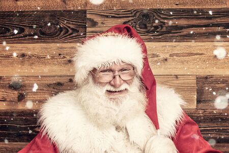 Smiling real Santa Claus in red cap posing on wooden wall, looking at camera. Merry Christmas and...
